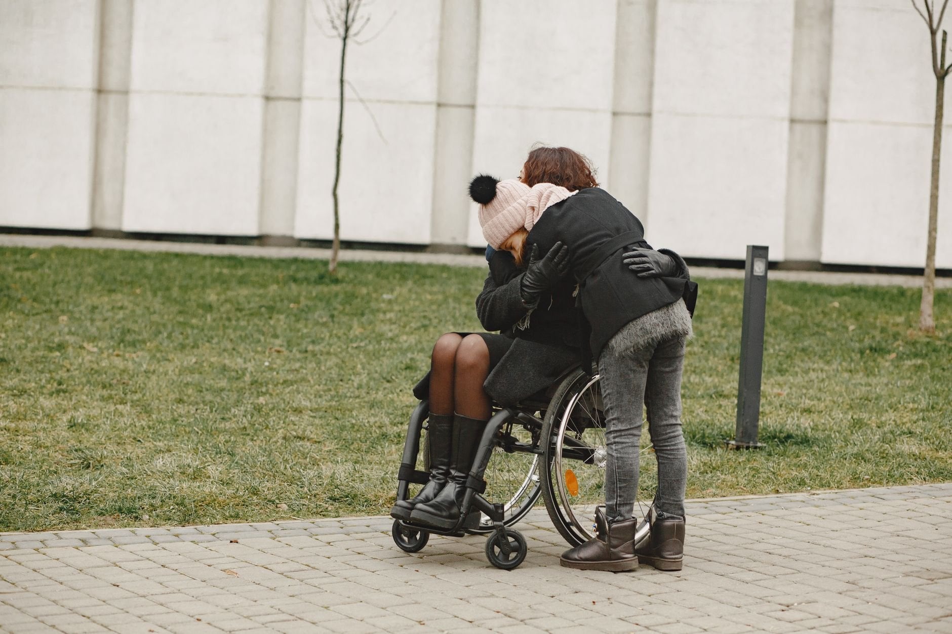 Two friends embrace warmly, one seated in a wheelchair, sharing affection outdoors.